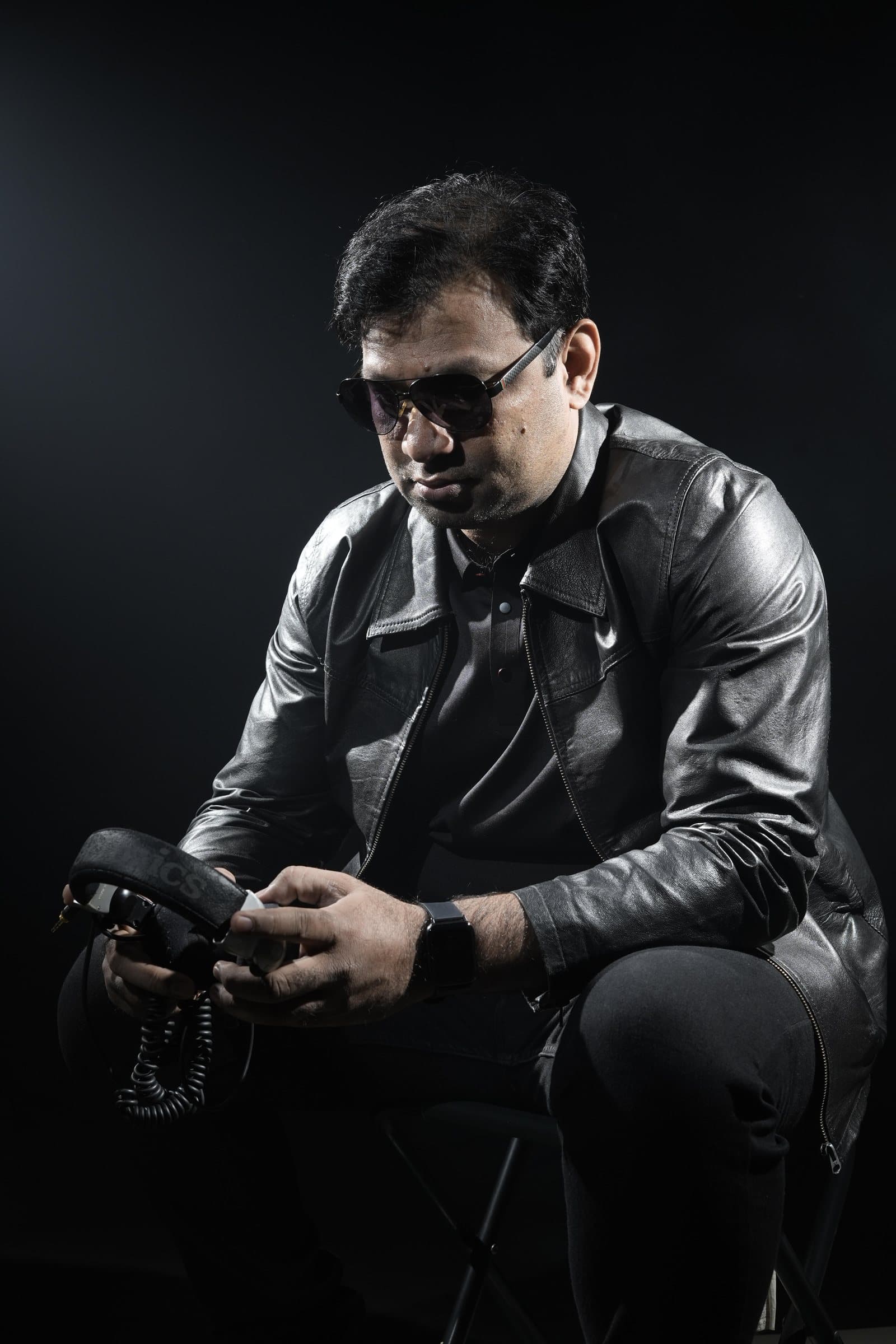 DJ Abdul crouched in a leather jacket, holding a pair of studio headphones